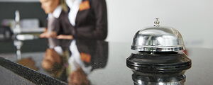 Hotelrezeption mit Fokus auf eine Klingel, im Hintergrund zwei Mitarbeiterinnen in Businesskleidung