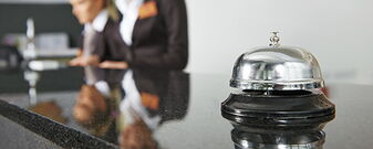 Hotelrezeption mit Fokus auf eine Klingel, im Hintergrund zwei Mitarbeiterinnen in Businesskleidung